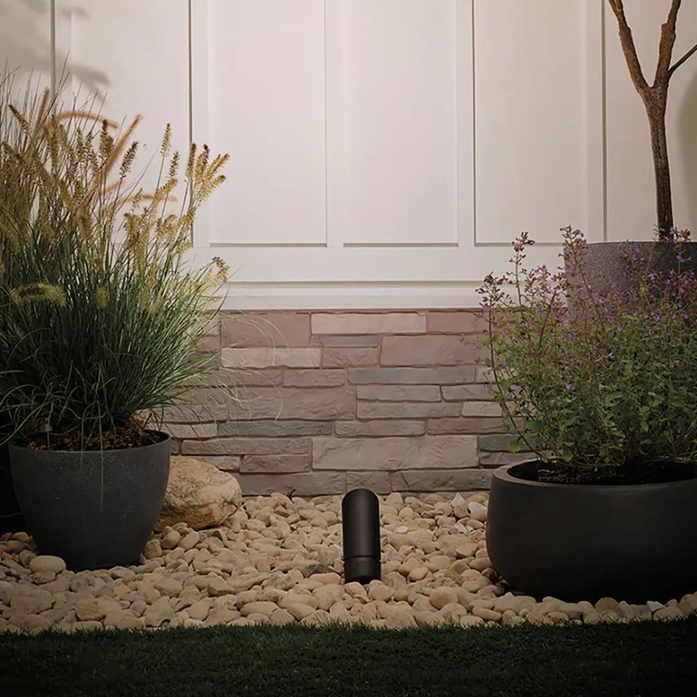 Black outdoor landscape LED accent light standing in gravel between potted plants